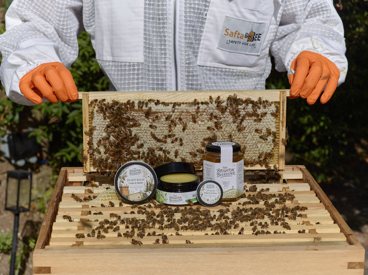 A beekeeper holding a frame of honey above The Brighton Beekeeper beeswax skincare on a cedar hive