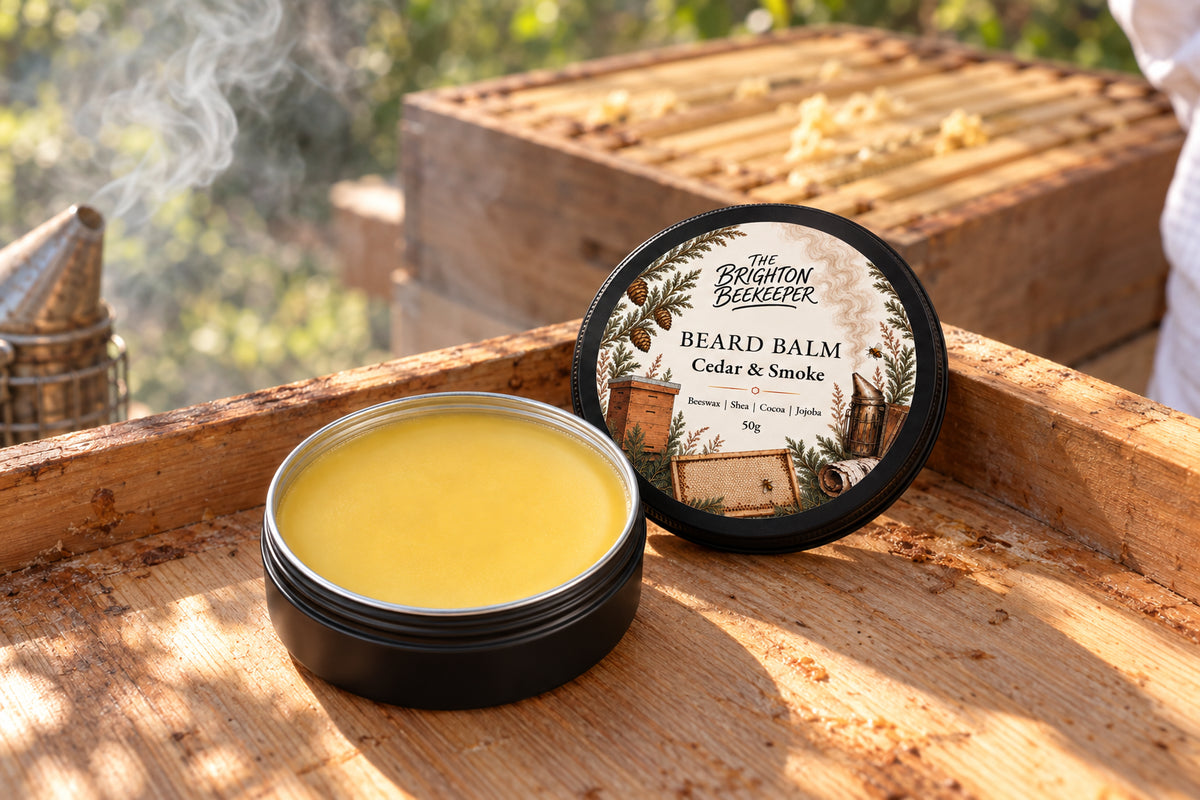 Open Cedar & Smoke Beard Balm on a wooden hive roof, with honeycomb frames and a beekeeper’s smoker behind