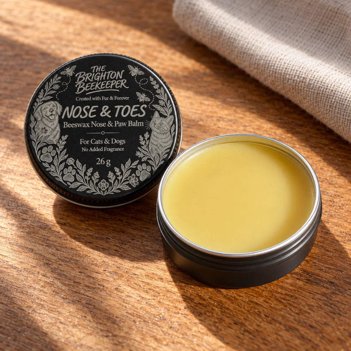 An open black Nose & Toes tin showing the smooth golden balm, with its silver-marked lid alongside